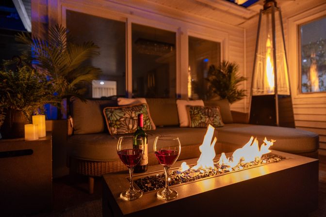 Cozy outdoor patio at night with two glasses of red wine and a bottle on a fire table, warm flames beside a cushioned wicker sofa with decorative pillows and potted palms