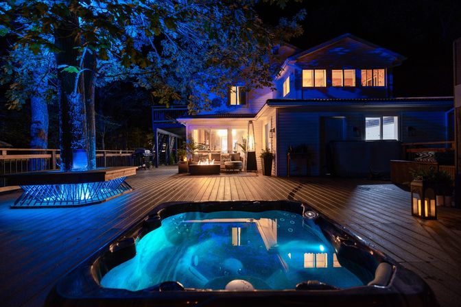 Nighttime wooded backyard deck with glowing hot tub in foreground, blue uplighting on trees, warm-lit modern two-story house, cozy outdoor fire pit lounge and lanterns on the patio.