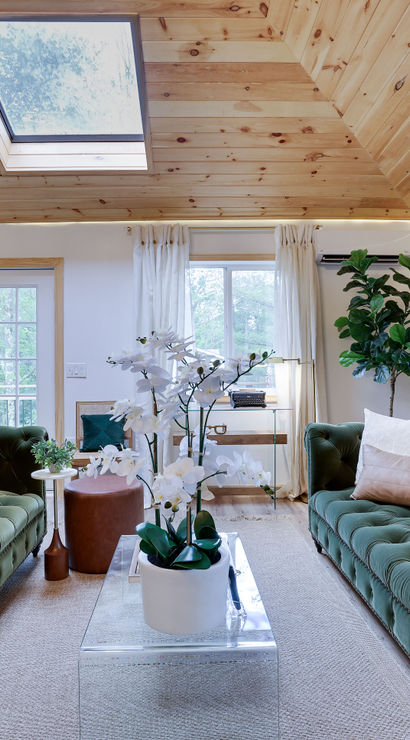 Bright cozy living room with twin green velvet tufted sofas, glass coffee table with white orchid centerpiece, wood-paneled vaulted ceiling with skylight, and French doors letting in natural light.