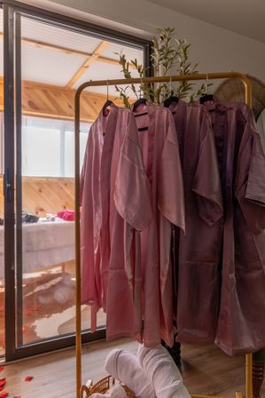 Pink satin robes hanging on a gold clothing rack in a cozy wood-paneled bedroom near a sliding glass door, with a towel basket and scattered rose petals on the floor.