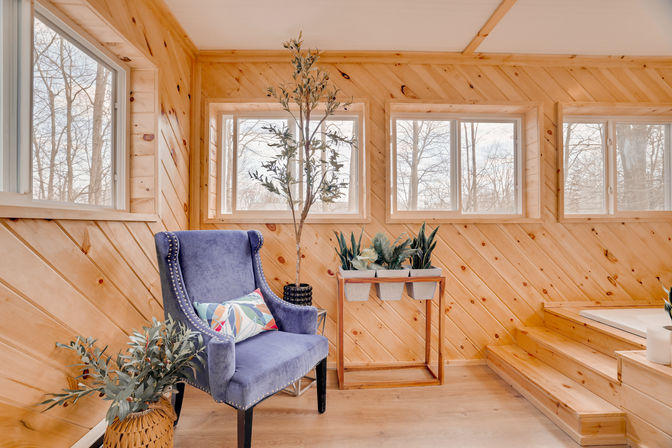 Cozy bright wood-paneled sunroom reading nook with a blue velvet wingback chair and colorful pillow, potted plants on a wooden stand, large windows and warm natural light.