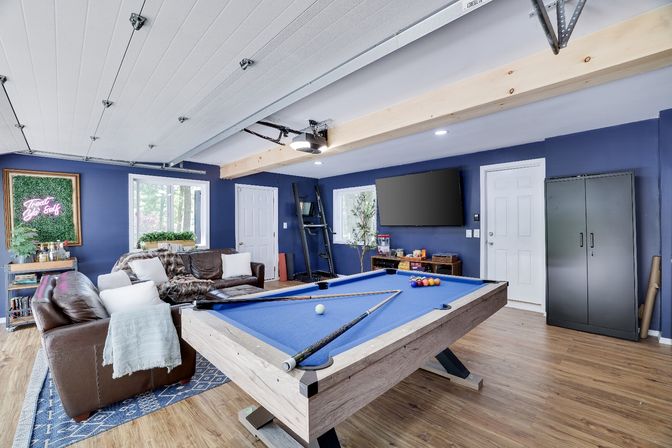 Game-ready modern rec room with a blue-felt pool table, leather sofas on a patterned rug, navy blue walls, hardwood floors, wall-mounted TV, neon wall art and storage cabinet