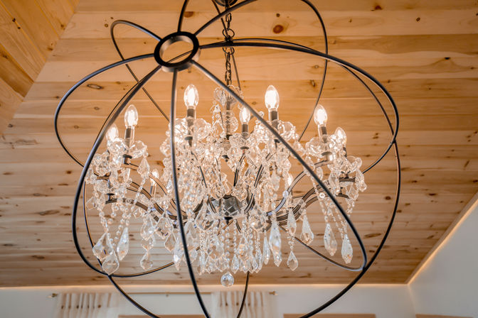 Sparkling crystal chandelier with teardrop prisms encased in a black metal orb, glowing beneath a warm pine wood ceiling in a rustic-modern interior