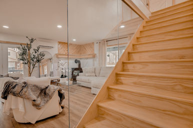 Cozy modern living room with light wood staircase, glass partition, neutral sectional, wood-burning stove and faux-fur throw