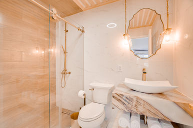 Bright modern bathroom with glass walk-in shower, gold fixtures, white marble walls, wooden slab vanity with vessel sink, hanging pendant lights and rolled towels.