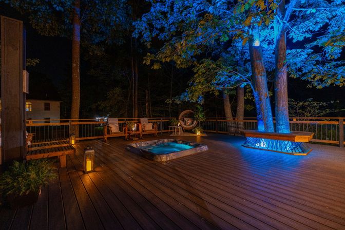 Nighttime backyard wood deck with a recessed hot tub, lantern-lit lounge chairs and a hanging wicker chair, bench built around trees highlighted by blue uplighting overlooking a wooded yard.
