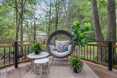 Cozy backyard deck with round wicker egg chair on legs, tropical-pattern throw pillow, nested white side tables and potted ferns, framed by wooden railing and leafy wooded yard