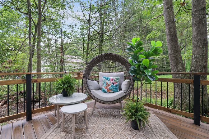 Cozy backyard deck with round wicker egg chair on legs, tropical-pattern throw pillow, nested white side tables and potted ferns, framed by wooden railing and leafy wooded yard