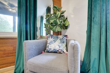 Cozy living room corner with a gray upholstered armchair, tropical patterned throw pillow with pink tassels, fiddle-leaf fig plant, teal velvet curtains and warm wood-paneled wall.