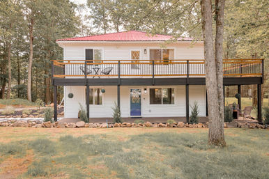 Charming two-story white country house with red metal roof, elevated wooden deck and blue front door, set on a grassy, tree-lined lot with stone-edged landscaping.