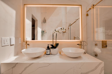 Spa-like modern bathroom vanity with double white vessel sinks on marble countertop, backlit mirror, gold fixtures, white orchid and glass shower