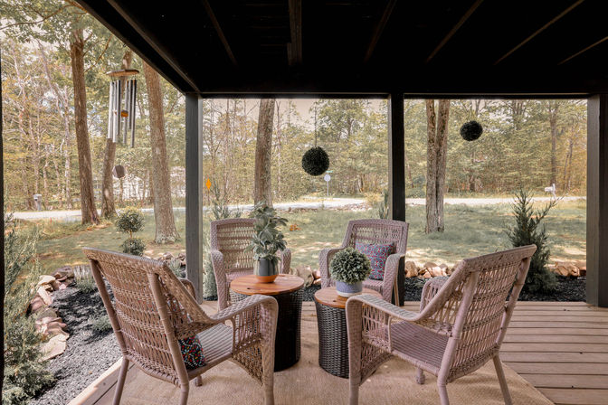 Cozy covered porch with four wicker chairs around two wood‑top tables, potted plants, hanging greenery and a wind chime, overlooking a tree‑lined yard and quiet country road.