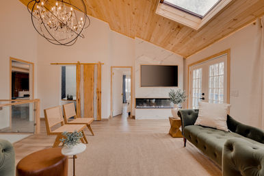 Light-filled modern farmhouse living room with vaulted pine ceiling and skylight, crystal chandelier, marble fireplace with mounted TV, green tufted sofa, natural wood sliding barn door, rattan chairs and French doors