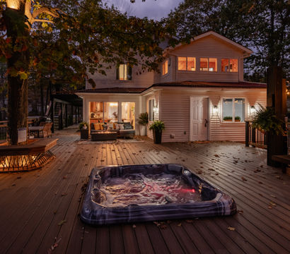 Evening backyard deck of a two-story suburban home in a wooded setting, bubbling hot tub on a wide wooden deck, cozy outdoor seating, fire pit, string lights and autumn leaves