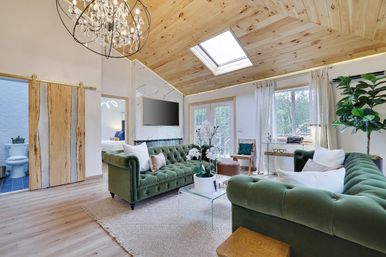 Sunlit modern farmhouse living room with vaulted wood ceiling and skylight, crystal chandelier, two green velvet tufted sofas around a glass coffee table with white orchids, sliding barn door and French doors to the outside.