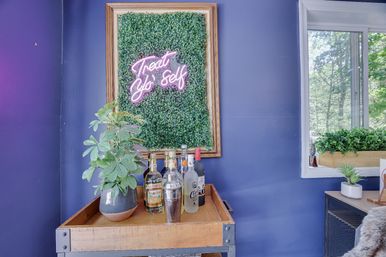 Indoor bar cart against a deep purple wall with framed boxwood panel and playful pink neon sign reading 'Treat Yo' Self', potted plant, liquor bottles and shaker, and a window planter with trees outside