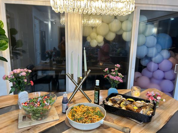Cozy home dining table setup with roasted chicken in a roasting pan, skillet of pasta, large salad bowl, olive oil, pink flower vases, lit candelabra and a pastel balloon garland reflected in the window — festive dinner spread.