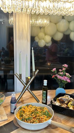 Cozy home dining table setup with roasted chicken in a roasting pan, skillet of pasta, large salad bowl, olive oil, pink flower vases, lit candelabra and a pastel balloon garland reflected in the window — festive dinner spread.