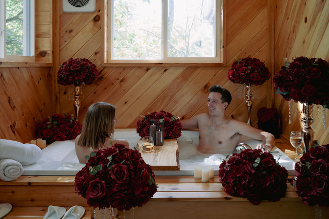 Romantic couple soaking in a bubble bath inside a rustic wood-paneled cabin bathroom, surrounded by deep-red rose bouquets, candlelight, wine glasses and a wooden bath caddy beneath a window.