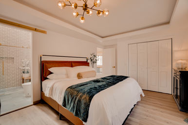 Bright modern bedroom with large upholstered bed, white linens and teal velvet throw, brass mid-century chandelier, wood floors, double closet doors and an open ensuite bathroom with patterned tile.