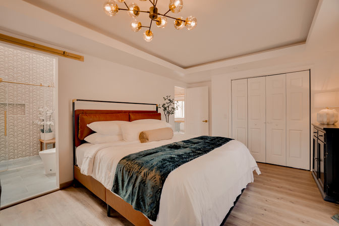 Bright modern bedroom with large upholstered bed, white linens and teal velvet throw, brass mid-century chandelier, wood floors, double closet doors and an open ensuite bathroom with patterned tile.