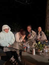 Four people bundled in faux-fur blankets on a backyard patio at night, laughing and sharing drinks around a table with water bottles, a soda can, cups and a small potted plant.