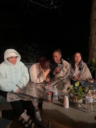 Four people bundled in faux-fur blankets on a backyard patio at night, laughing and sharing drinks around a table with water bottles, a soda can, cups and a small potted plant.
