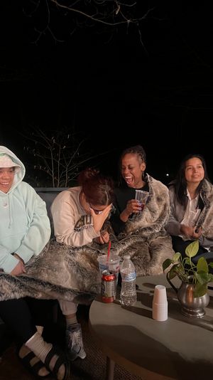 Four people bundled in faux-fur blankets on a backyard patio at night, laughing and sharing drinks around a table with water bottles, a soda can, cups and a small potted plant.
