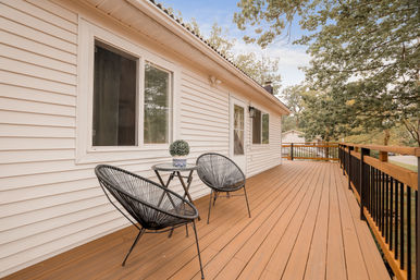 Cozy wooden backyard deck with two black woven patio chairs and a small round table with a potted plant against a white-sided house, overlooking a tree-lined yard.