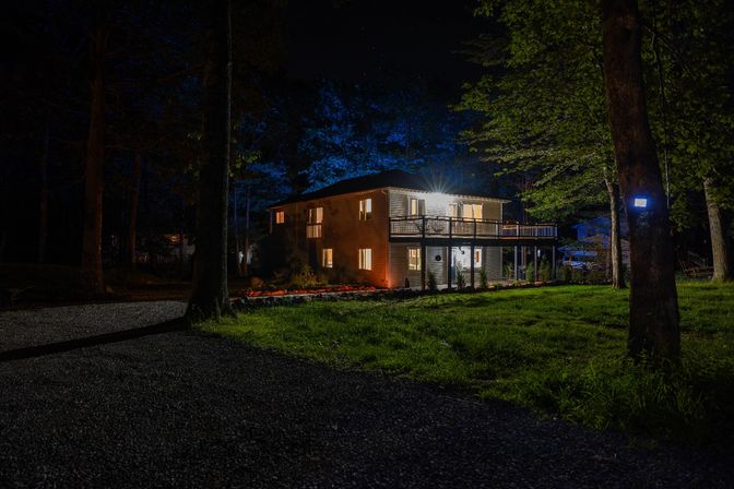 Cozy two-story house at night in a wooded yard, warm interior lights and a lit second-floor deck, gravel driveway, trees with blue landscape uplighting and outdoor security light