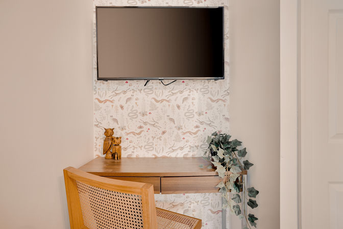 Cozy home office nook with wall-mounted flat-screen TV above a wooden desk, cane-back chair, carved owl figurines and trailing ivy against whimsical botanical wallpaper.