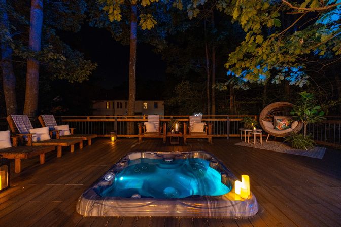 Cozy nighttime backyard deck with glowing blue hot tub on a wooden patio, cushioned lounge chairs, a wicker egg chair, lantern candles and tree canopy