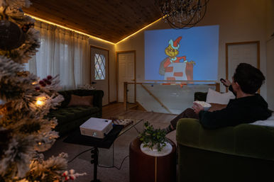 Cozy holiday living room and home theater—lit Christmas tree and warm LED trim, person on a green sofa with popcorn and remote watching a festive cartoon projected on the wall from a tabletop projector.