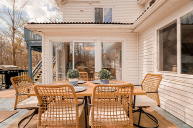 Outdoor backyard deck dining area at a white-sided suburban house: wooden table with woven wicker chairs, potted topiary centerpieces, sliding glass doors and a grill nearby.