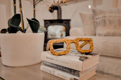 Quirky carved wooden eyeglass sculpture on a stack of books on a glass coffee table beside a potted orchid, set in a cozy neutral living room with a wood-burning stove in the background.
