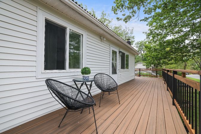 Spacious backyard wooden deck with brown planks, black woven patio chairs and small round table with potted plant, attached to a white-sided house and overlooking leafy suburban trees and railing.
