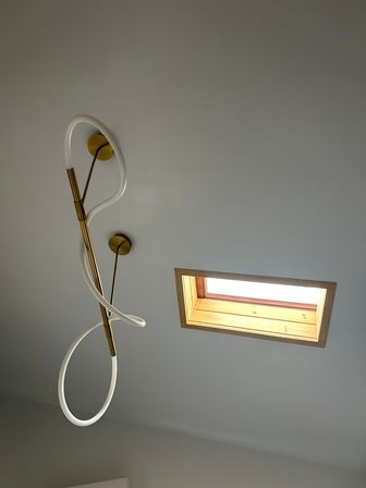 Modern twisted LED pendant light with brass fittings on a pale gray interior ceiling beside a rectangular wooden-framed skylight letting in natural light.