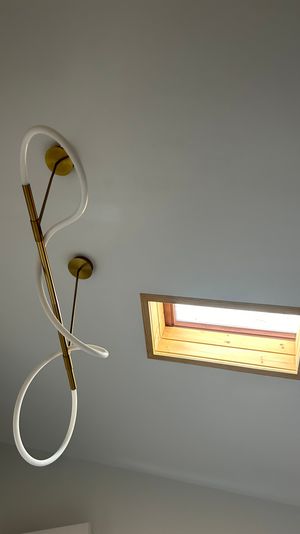 Modern twisted LED pendant light with brass fittings on a pale gray interior ceiling beside a rectangular wooden-framed skylight letting in natural light.