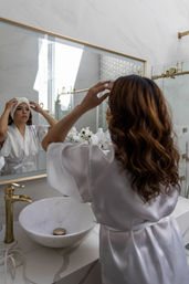 Woman in a white satin robe adjusting a towel in a bright modern marble bathroom with gold fixtures, round vessel sink, and white orchids reflected in the mirror.