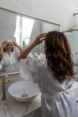Woman in a white satin robe adjusting a towel in a bright modern marble bathroom with gold fixtures, round vessel sink, and white orchids reflected in the mirror.