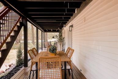 Cozy covered backyard porch dining area with wooden table and woven wicker chairs under black-beamed deck, outdoor staircase and leafy yard view