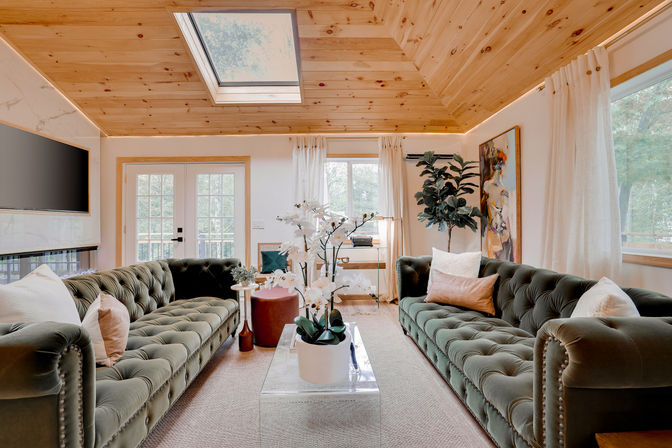 Sunny cozy living room with vaulted pine ceiling and skylight, twin tufted green velvet sofas facing a clear acrylic coffee table topped with white orchids, French doors and large windows bringing in natural light
