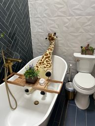 Plush giraffe lounging in a white freestanding bathtub with a wooden bath tray, gold faucet, dark herringbone and textured gray wall tiles, and a toilet with succulent planter in a modern bathroom.