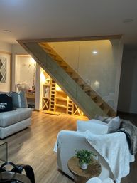 Cozy modern home living room with warm-lit wooden staircase and glass railing, built-in under-stair shelving and wine cubbies, hardwood floors, plush white armchair and small wooden side table with potted plant.