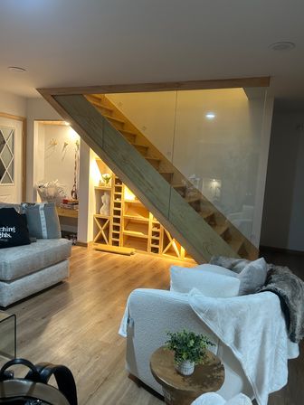 Cozy modern home living room with warm-lit wooden staircase and glass railing, built-in under-stair shelving and wine cubbies, hardwood floors, plush white armchair and small wooden side table with potted plant.