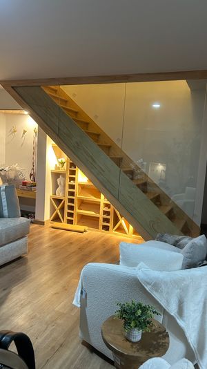 Cozy modern home living room with warm-lit wooden staircase and glass railing, built-in under-stair shelving and wine cubbies, hardwood floors, plush white armchair and small wooden side table with potted plant.