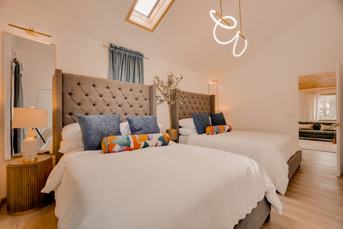 Bright modern bedroom interior with two upholstered tufted-headboard beds, white linens, blue throw pillows and colorful bolster cushions, skylight and sculptural pendant light, wooden floor and bedside lamp.