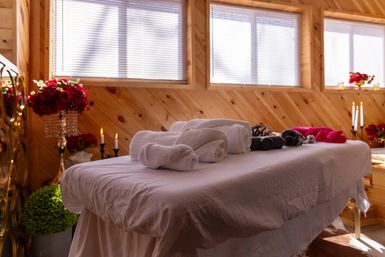 Cozy wood-paneled spa treatment room with a massage table draped in white linens, rolled towels, lit candles and red floral arrangements beneath sunlit windows.