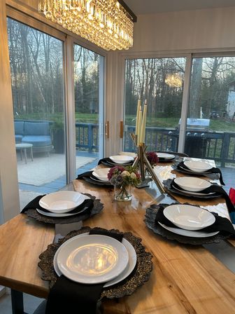 Wooden dining table set for six with white plates, black napkins and textured chargers, floral centerpiece and tall candles under a sparkling crystal chandelier by sliding glass doors overlooking a wooded backyard deck with a grill.
