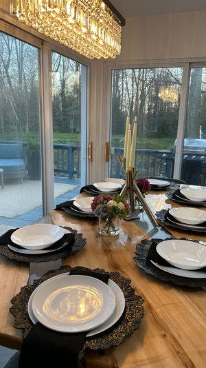 Wooden dining table set for six with white plates, black napkins and textured chargers, floral centerpiece and tall candles under a sparkling crystal chandelier by sliding glass doors overlooking a wooded backyard deck with a grill.